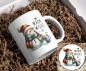 Preview: Weihnachtstasse mit Schneemann und ‚Warm Wishes‘-Motiv sowie passendem Untersetzer: Ein gemütlicher Schneemann mit rot-grün kariertem Schal und roter Mütze hält eine Tasse heißen Kakao mit Zuckerstange. Über dem Motiv steht der Schriftzug ‚Warm Wishes‘ ve